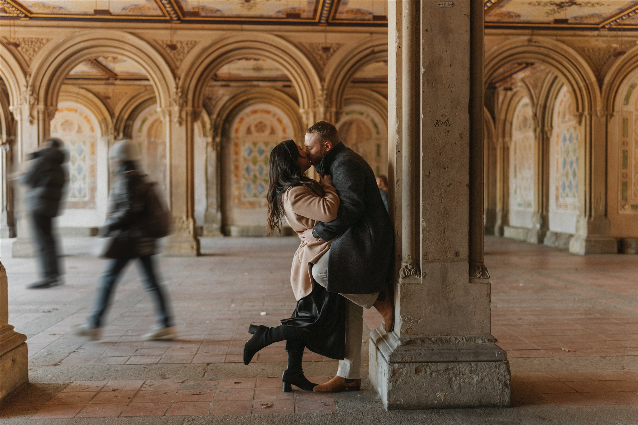 Engaged couple kissing while holding onto each other's long coats in Central Park's Bethesda Terrace as strangers walk by for their NYC Central Park Engagement